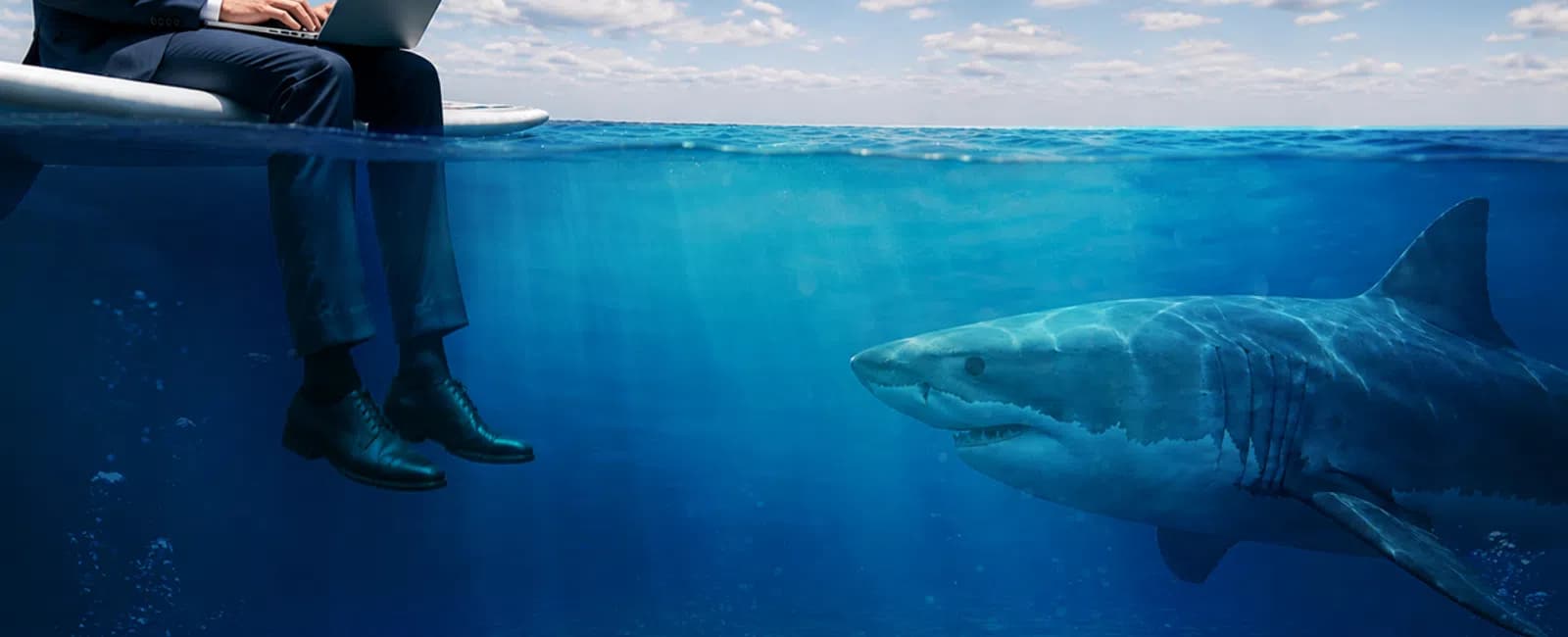 Businessman working on laptop while sitting above water with a shark swimming below him in clear ocean.