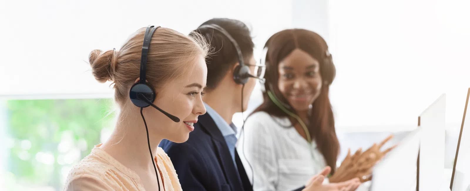 Diverse customer support team wearing headsets working on computers and communicating in a bright office.
