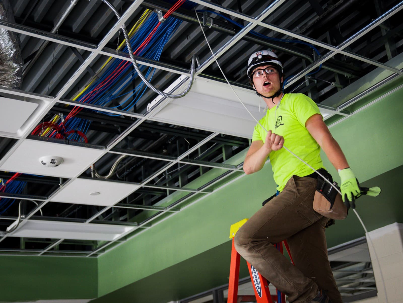 A Quantum technician in a hard hat and safety glasses on a ladder, pulling network cable through a suspended ceiling grid in a commercial building.