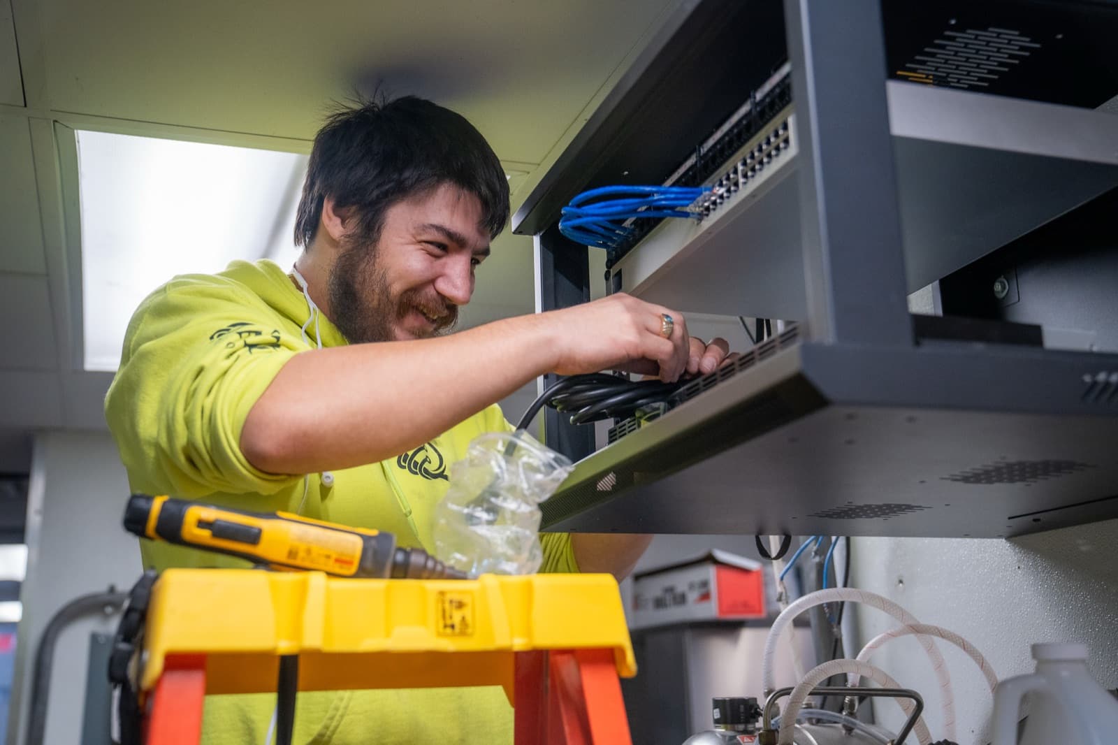 A Quantum technician on a stepladder, reaching into a wall-mounted network cabinet to connect cabling at a client site.